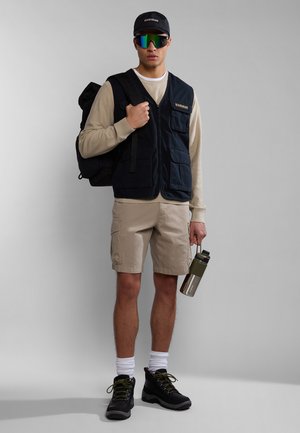 Gilet blu con molte tasche sopra un maglione beige, pantaloni cargo color sabbia, stivali neri stringati e una borraccia verde. Occhiali da sole riflettenti.