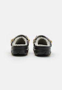 Zapatos negros tipo mocasín con forro de felpa beige, respaldos acolchados y correas ajustables amarillas; suelas de goma texturizada para mayor tracción.