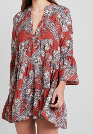 Nainen, jolla on päällään löysä, punainen kuvioitu mekko, jossa on kolme neljäsosaa hihasta ja syvä V-aukko, yksinkertaisen harmaan taustan edessä.