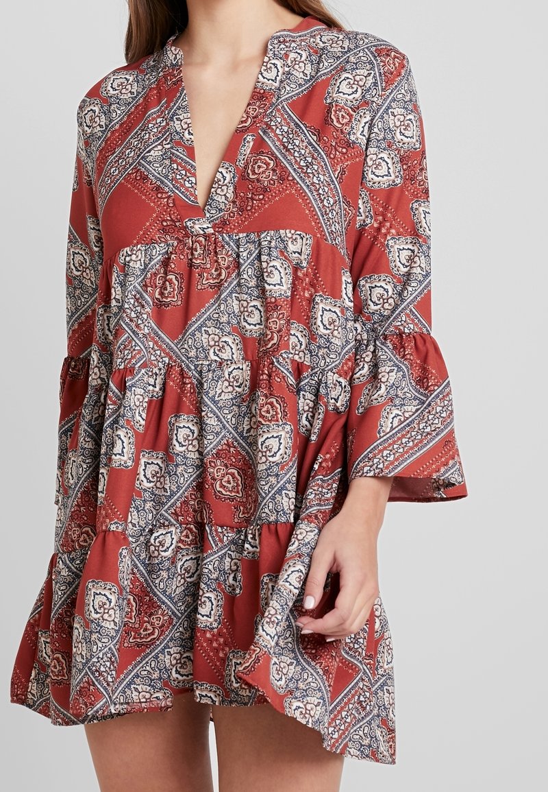 Femme portant une robe ample rouge à motifs avec des manches cloche trois-quarts et un décolleté en V profond, sur un fond gris uni.