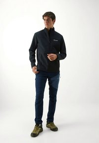 Sort softshelljakke med høy krage, hel glidelås og logo. Mørkeblå bukser med sidelomme. Olivengrønne tursko.