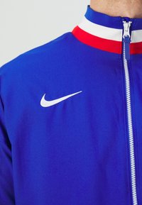 Nike zip-jakke i livlig blå med hvidt logo, udstyret med en ribbet krave i rød og hvid samt glat, letvægtsstof.