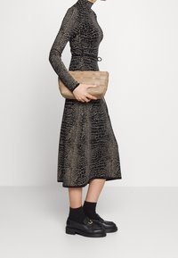 Robe noire avec un motif texture crocodile et des manches longues, assortie à une pochette tissée beige et des mocassins noirs avec des détails en métal doré.