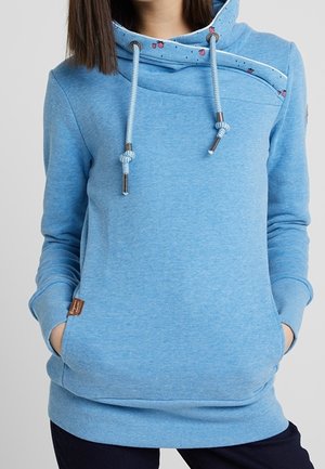 Femme portant un sweat à capuche bleu clair avec cordons, col croisé unique avec petites impressions de coccinelles, et les mains dans les poches avant.