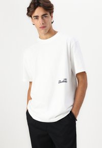 T-shirt de algodão branca com decote redondo e mangas curtas. Apresenta um logo preto no lado esquerdo do peito. Usada com calças pretas.