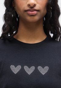 Μαύρο t-shirt με τρία σχήματα καρδιάς από μικρές, ασημένιες στρας. Στρογγυλή λαιμόκοψη με απαλή υφή. Απλός και μοντέρνος σχεδιασμός.