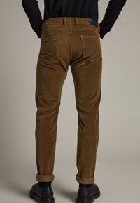 Brown corduroy trousers with a fitted design, featuring back pockets, a rolled cuff, and a textured fabric. Black shoes visible at the bottom.