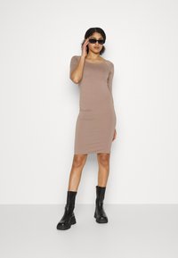 Vestido bodycon em tecido castanho claro com decote quadrado, mangas curtas e comprimento até à metade da coxa. Combinado com botas pretas robustas e óculos de sol.
