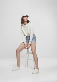 Hvit hettegenser med lange ermer, lyseblå denim-shorts og hvite joggesko, stylet med en grå caps, sittende på en hvit krakk.