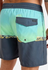 Męskie kąpielówki z gradientem od turkusu do granatu, elastycznym pasem, boczną kieszenią z klapką oraz żółtym logo na dolnym brzegu.