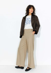 Veste en imprimé léopard marron, t-shirt graphique blanc, pantalons larges beiges, et chaussures plates noires avec des accents ronds.