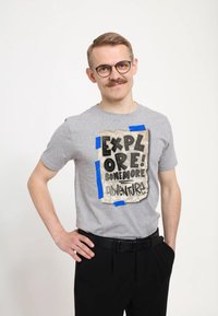 Šedé tričko s texturovaným designem, na kterém jsou vytištěna slova "PROZKOUMEJ! JEŠTĚ VÍCE DOBRODRUŽSTVÍ!" a doplněna modrou páskou.