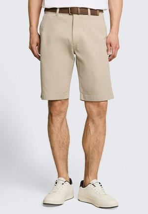 Man met beige knielange korte broek, bruine gevlochten riem en witte sneakers, staand tegen een effen lichte achtergrond.