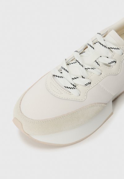 Adidași albi cu vârf din suede texturat și partea superioară din țesătură netedă. Au șiret alb cu accente negre și o talpă căptușită.