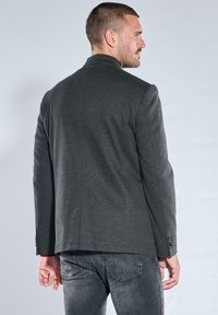 Gray blazer in a structured design with a high collar, two back vents, and button accents on the cuffs, worn over gray jeans.