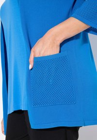 Blå tunika med en mjuk yta och en kontrasterande meshficka. Fickan har en perforerad design som ökar andningsbarheten.