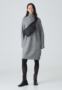 Übergroßes graues gestricktes Kleid mit hohem Kragen, getragen mit schwarzen Stiefeletten und einer schwarzen Umhängetasche. Der Hintergrund ist schlicht.