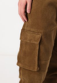 Braune Cord-Hosen mit einer strukturierten Oberfläche, ausgestattet mit einer Seitentasche, sichtbaren Nähten und einem taillierten Design entlang des Beins.
