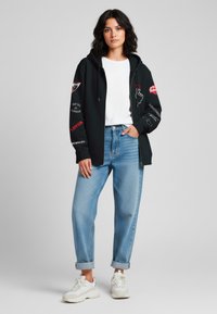 Felpa nera con zip e stampe grafiche, abbinata a jeans azzurri con risvolti e sneakers bianche. La felpa presenta vari design colorati.