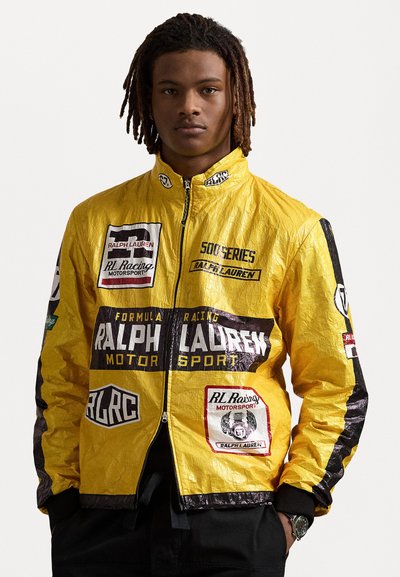 Geel-zwart racjacket met een glanzende, kreukelige textuur. Bevat meerdere logo's en gedurfde typografie. Voorzijde met rits en opstaande kraag.