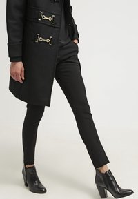 Manteau noir avec des accessoires dorés, associé à un pantalon ajusté noir et des bottines à talons, mettant en valeur des lignes épurées et une texture lisse.