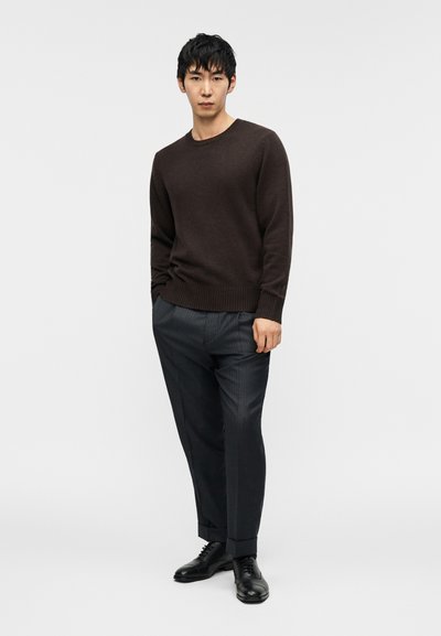 Σκούπε καφέ μάλλινο πουλόβερ με ριμπ τελείωμα, συνδυασμένο με γκρι ριγέ παντελόνια. Μαύρα δερμάτινα παπούτσια ολοκληρώνουν το σύνολο. Το μοντέλο στέκεται σε ουδέτερη στάση.