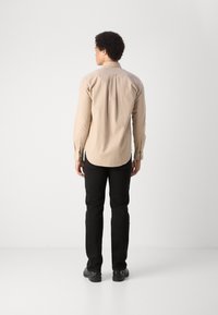 Camicia a maniche lunghe color beige chiaro con orlo curvo, polsini con bottoni e piega sul retro, abbinata a pantaloni neri slim-fit e scarpe nere.