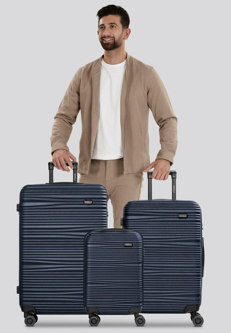 Homme portant une veste beige et un pantalon, se tenant derrière trois valises en bleu marine à nervures avec des poignées extensibles, sur fond gris.