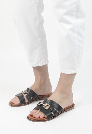 Schwarze Ledersandalen mit texturierter Oberfläche, einem goldfarbenen Metallakzent und einer flachen braunen Sohle.