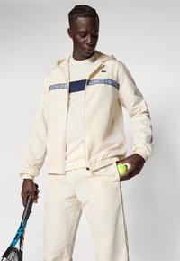 Krema zip-up jopica z mornarsko modrim akcentom, bela majica in kremne hlače. Tenis raketa v roki, zelena tenis žogica v žepu.