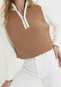 Bruin en crème gebreide pullover met een hoge kraag en ritssluiting, voorzien van fijne ribbels en contrasterende kleurvlakken. Gecombineerd met witte jeans.