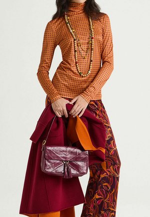 Maglia arancione a maniche lunghe con pois rossi, abbinata a un cappotto color borgogna, pantaloni vivaci con fantasie, e una piccola borsa borgogna con nappine.