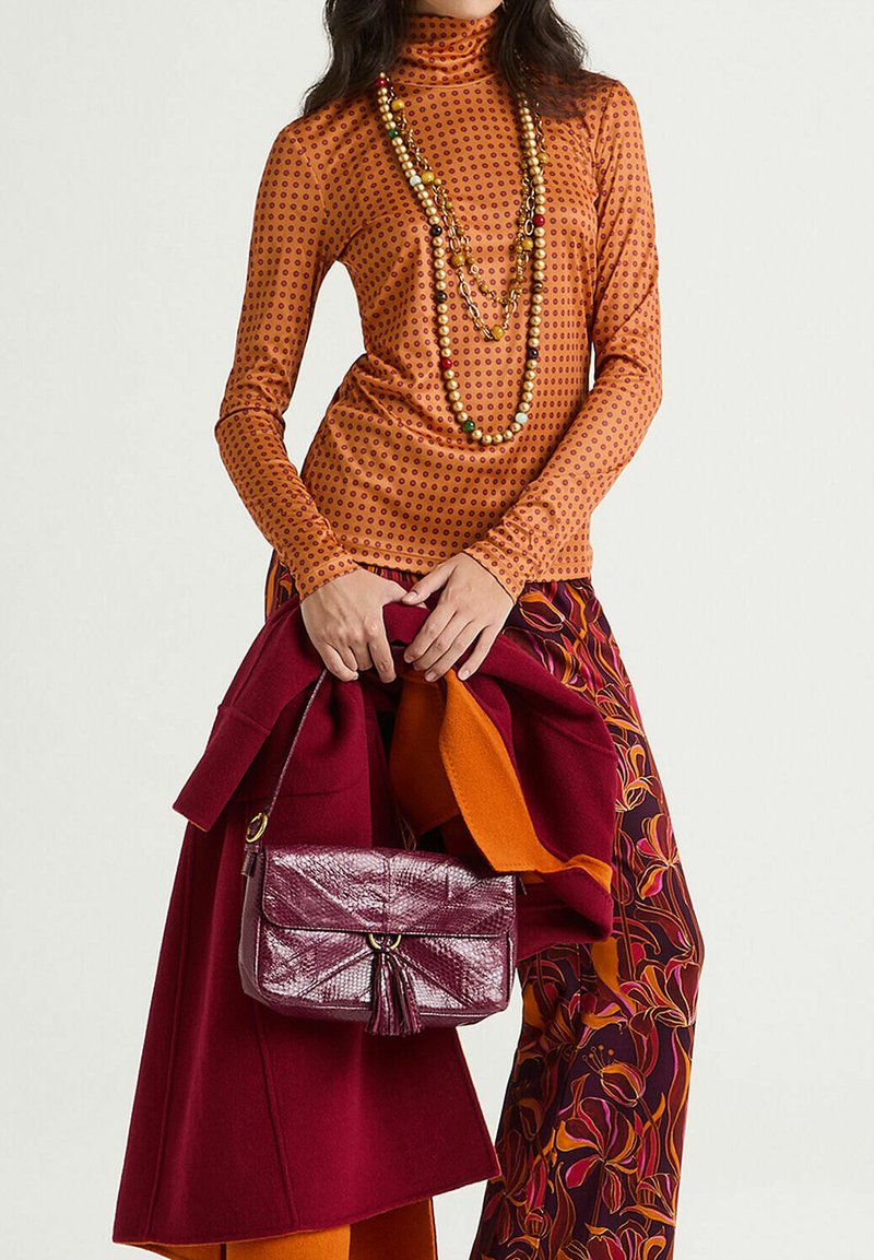 Haut à manches longues orange à pois rouges, superposé d'un manteau bordeaux, pantalon aux motifs vibrants, et un petit sac à main bordeaux avec un pompon.