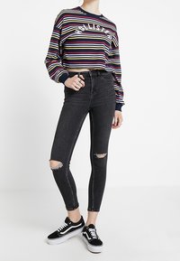 Kvinna som bär en randig långärmad croppad topp, rivna svarta skinny jeans och svart-vita sneakers stående mot en vit bakgrund.