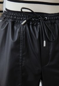 Pantalones cortos de imitación de cuero negro con una cinturilla elástica y cordón ajustable. Cuenta con bolsillos laterales y una textura suave. Acentos en hardware plateado.