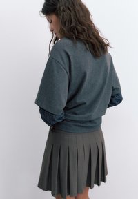 Kobieta z długimi, falowanymi włosami, ubrana w szarą, oversize'ową bluzę na niebieską koszulę w kratę i ciemną plisowaną spódnicę, zwrócona bokiem.