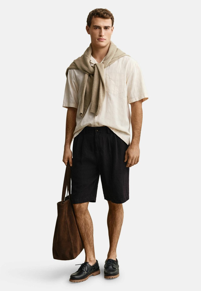 Hombre con camisa beige de manga corta y un suéter marrón claro sobre los hombros, pantalones cortos negros, zapatos negros y lleva un bolso tote marrón.