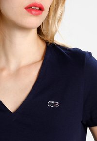 Granatowa koszulka V-neck z krótkim rękawem, z małym haftowanym logo krokodyla w białym i czerwonym kolorze na lewej części klatki piersiowej.