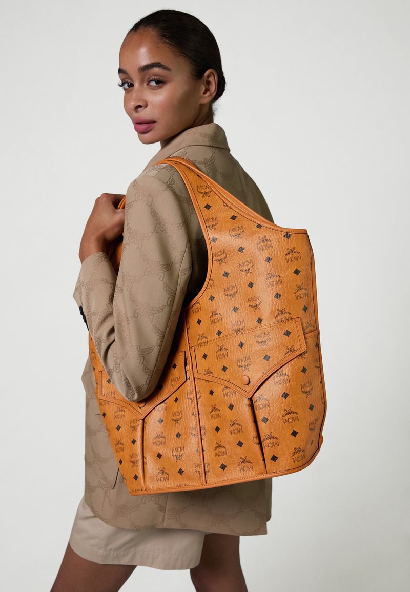 Woman wearing a patterned beige blazer and shorts holds large orange leather MCM tote bag with black logo print over shoulder.