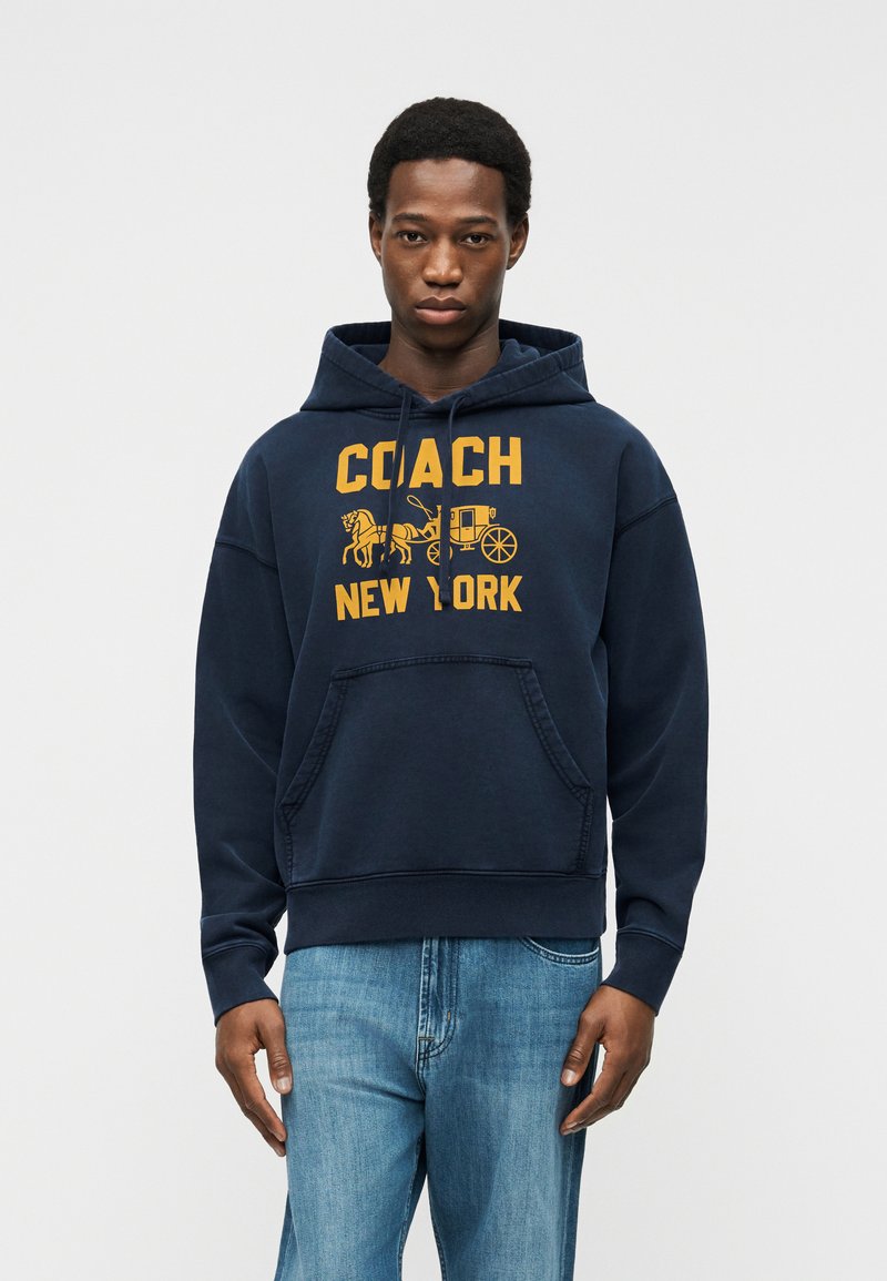 Mann trägt einen marineblauen Kapuzenpullover mit gelbem "Coach New York"-Schriftzug und Pferdekutschen-Grafik, kombiniert mit hellblauen Jeans vor einem schlichten Hintergrund.