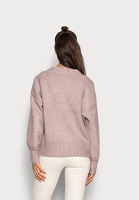 Světle růžový pletený svetr s robustní strukturou, spadlými rameny a žebrovaným lemem. Nesený přes bílé kalhoty, ukazující zadní pohled.