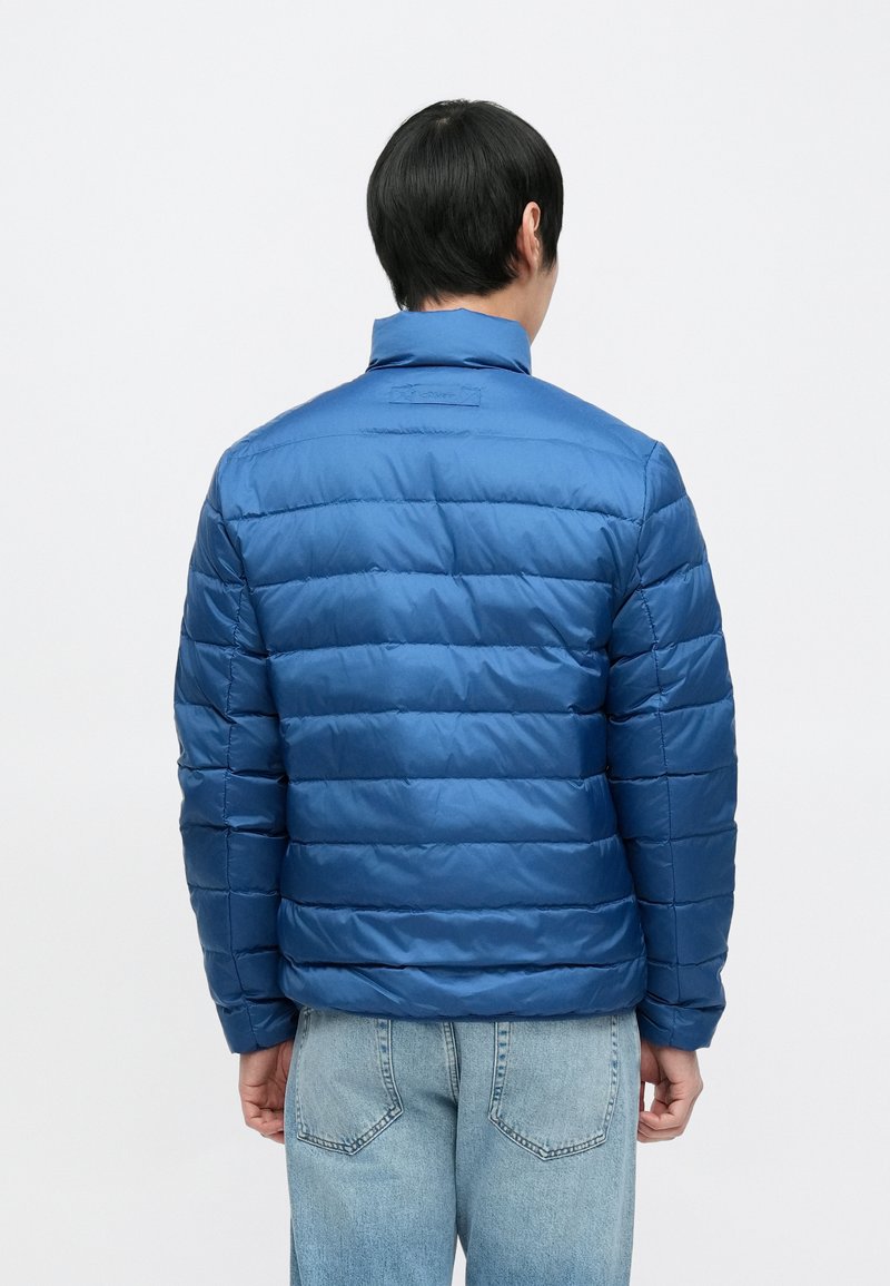 Blue puffer jacket with a quilted design and stand-up collar, featuring a smooth, shiny texture. Worn over light blue jeans.