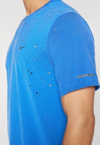 Sinine sportlik T-särk, millel on kulunud tekstuur mitme väikese auguga, musta Nike logo ja helendava halli triibuga varrukal.