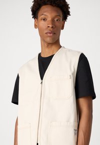 Beige Reißverschluss-Weste mit zwei Vordertaschen, aus strukturiertem Stoff. Trägt über einem schwarzen T-Shirt, das kontrastrierende Nähte und ein Etikett aufweist.