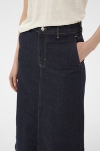Femme portant une jupe en denim bleu foncé avec des poches frontales et un haut blanc semi-transparent, la main posée dans la poche de la jupe.