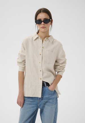 Vrouw draagt een beige overhemd met knopen, blauwe spijkerbroek, zwarte riem en zwarte cat-eye zonnebril, staat met één hand in haar zak.