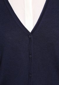 Cardigan bleu marine avec un col en V, tissu en tricot texturé, et six boutons assortis sur le devant, porté sur une chemise blanche.