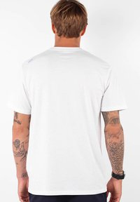 Camiseta de algodón blanca con mangas cortas, corte estándar. Presenta un pequeño detalle de logo en la parte superior de la espalda y tatuajes visibles en el brazo.