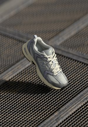Chaussure de course New Balance grise et blanche sur une surface en grille métallique avec motif texturé, éclairée par la lumière naturelle.