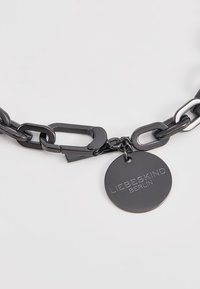 Bracelet en chaîne noire avec de grands maillons en métal, un fermoir et une étiquette ronde gravée "LIEBESKIND BERLIN". Texture lisse et finition brillante.